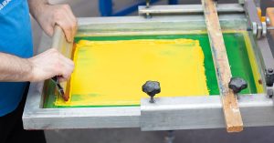 Professional demonstrating screen printing advantages using squeegee to apply bright yellow ink through mesh screen, showing precise technique and vibrant color saturation