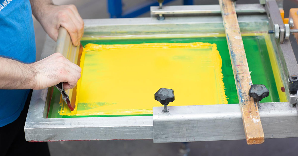 Professional demonstrating screen printing advantages using squeegee to apply bright yellow ink through mesh screen, showing precise technique and vibrant color saturation