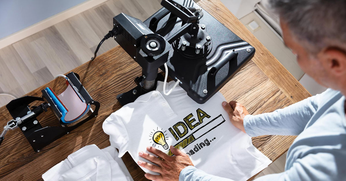 Professional heat transfer printing workspace showing heat press machine, cutting plotter, and white t-shirt with "IDEA" heat transfer vinyl design, illustrating what is heat transfer printing equipment and process