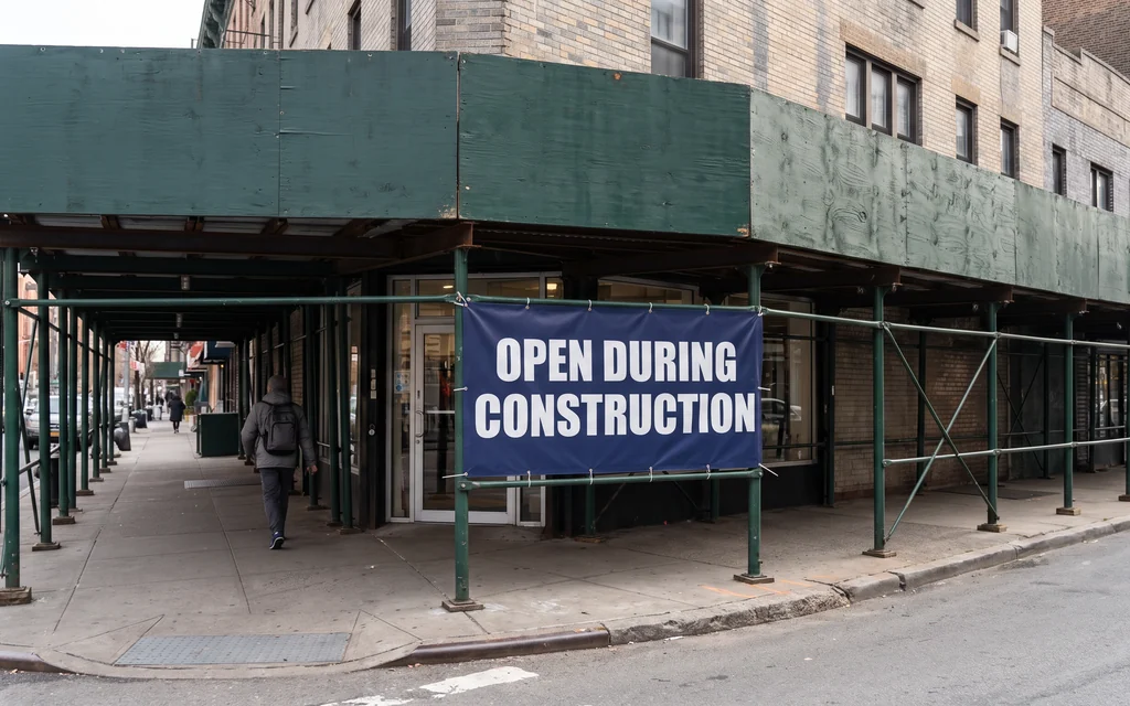 Open During Construction banner fastened to a green NYC sidewalk shed directly in front of the shop it hides