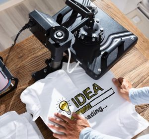 Professional heat transfer printing workspace showing heat press machine, cutting plotter, and white t-shirt with "IDEA" heat transfer vinyl design, illustrating what is heat transfer printing equipment and process