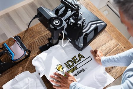 Professional heat transfer printing workspace showing heat press machine, cutting plotter, and white t-shirt with "IDEA" heat transfer vinyl design, illustrating what is heat transfer printing equipment and process