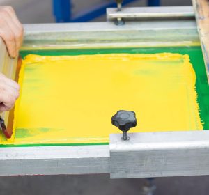 Professional demonstrating screen printing advantages using squeegee to apply bright yellow ink through mesh screen, showing precise technique and vibrant color saturation