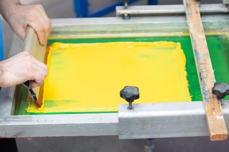 Professional demonstrating screen printing advantages using squeegee to apply bright yellow ink through mesh screen, showing precise technique and vibrant color saturation
